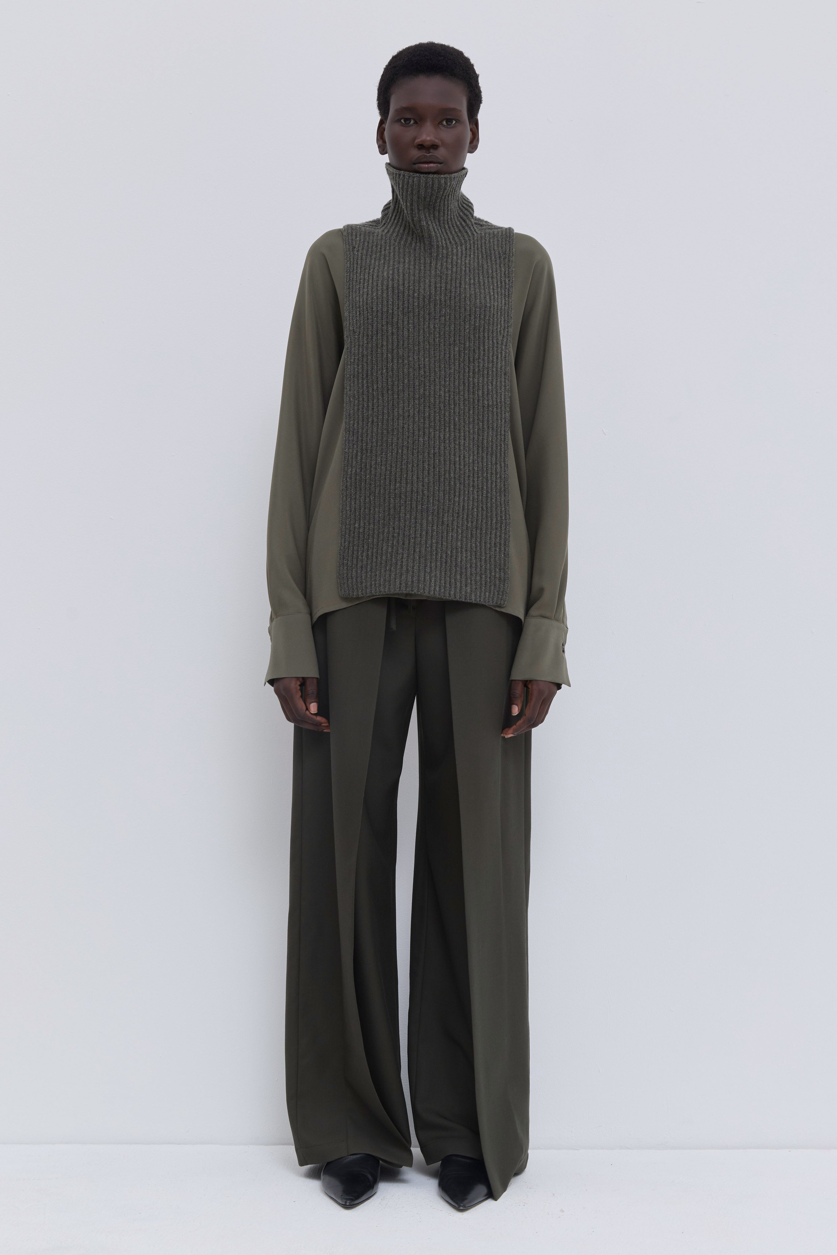 Forest melange ribbed merino wool and cashmere knit bib with oversized turtleneck, layered over an olive blouse and tailored wide-leg trousers.