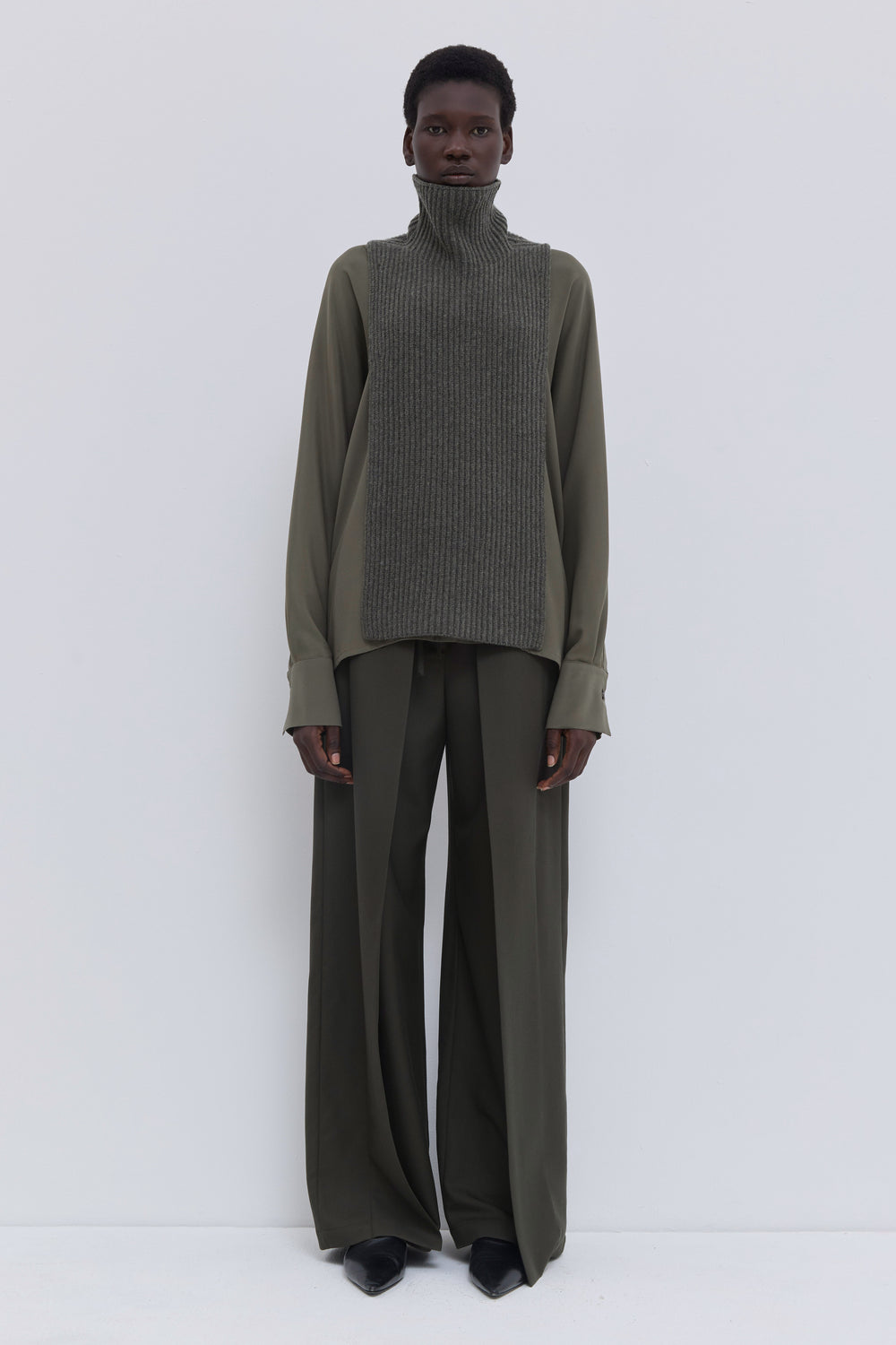 Forest melange ribbed merino wool and cashmere knit bib with oversized turtleneck, layered over an olive blouse and tailored wide-leg trousers.