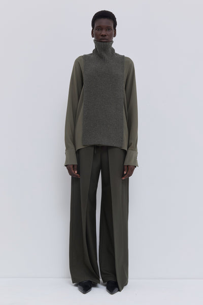 Forest melange ribbed merino wool and cashmere knit bib with oversized turtleneck, layered over an olive blouse and tailored wide-leg trousers.