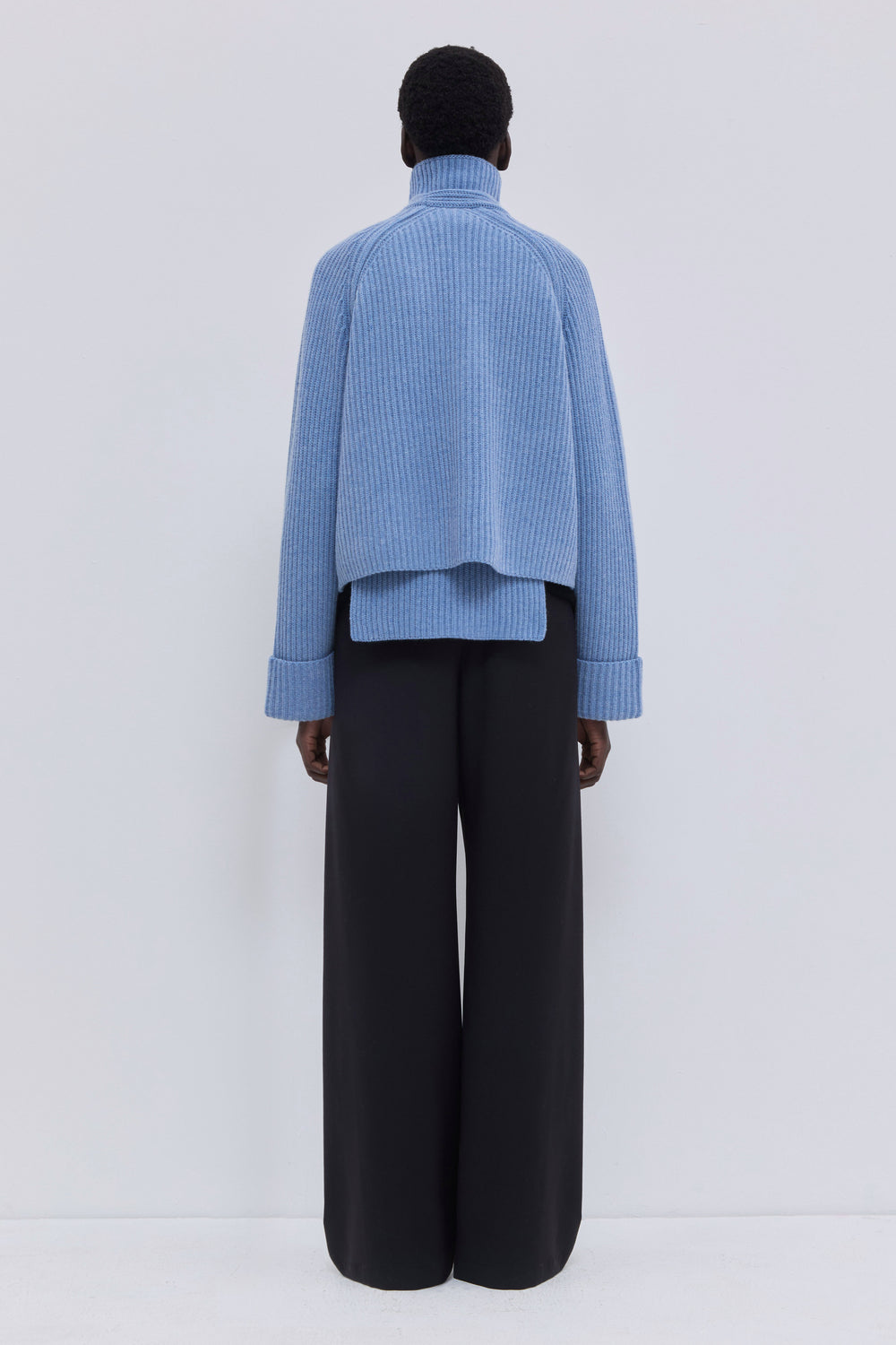 Back view of mirage blue ribbed merino wool and cashmere knit bib with elongated silhouette and oversized turtleneck.